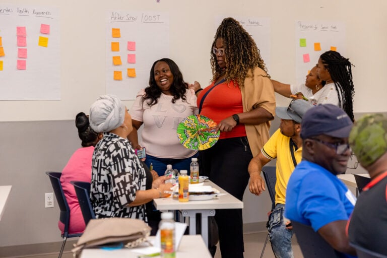 a small group of women huddle at a community meeting in front of a board with sticky-note ideas