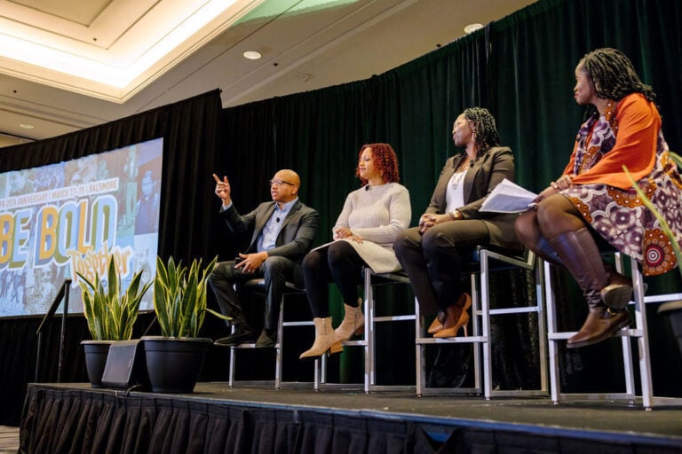 4 people on stools presenting at a conference with a "Be Bold" poster on a screen